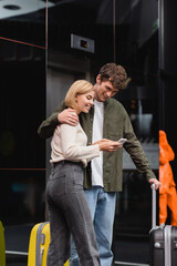 Obraz premium smiling woman showing mobile phone to boyfriend embracing her near baggage in hotel lobby.