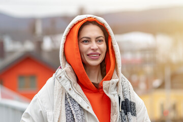 Street portrait of a smiling woman 25-30 years old in a white jacket on a bridge on a blurry background of city houses, color illumination.