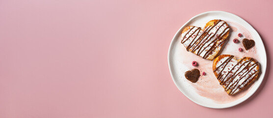 Valentine's Day wide banner with copy space. Heart-shaped French toast with cream, chocolate, and freeze-dried raspberry crumbles, on a ceramic plate against a light pink background.