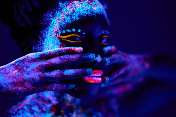 beautiful young black female with fluorescent prints on face, unusual prints, body art. neon lights, uv ray, luminescence concept. model touching face, covering. close-up portrait