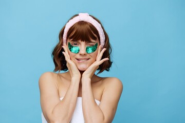 a happy woman stands on a blue background with colored patches on her eyes and holds her face with her hands while biting her lower lip