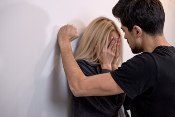 family violence. aggressive and nervous bearded guy arguing with crying blonde wife at home, caucasian woman is standing with back against the wall at home, suffering from domestic violence