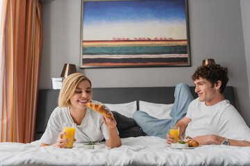 pleased couple having breakfast with orange juice and croissants in modern hotel bedroom.
