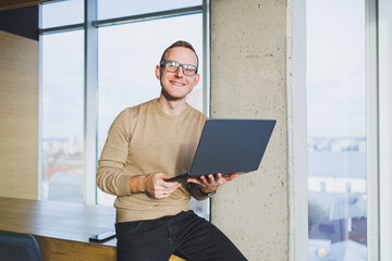 A businessman in glasses and stylish clothes drinks coffee in a modern workspace. A male freelancer with a laptop works remotely