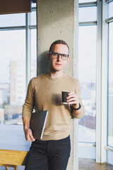 A businessman in glasses and stylish clothes drinks coffee in a modern workspace. A male freelancer with a laptop works remotely