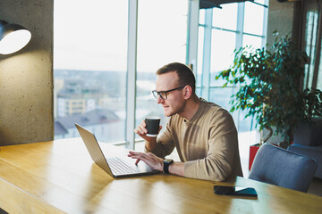 Young freelancer man in glasses sitting at a wooden table with a laptop and coffee while working on a remote project in a modern workspace.