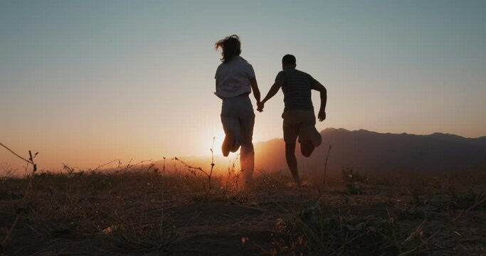 Back view of love couple silhouette running into the sunset. Happy young man and woman running holding hands, in nature 4k slowmotion