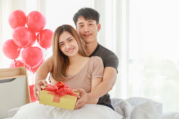 Couple and Valentine Day Concept. Asian smiling female and male holding and give a giftbox on white blanket on bed in bedroom with mini red heart shape pillow.