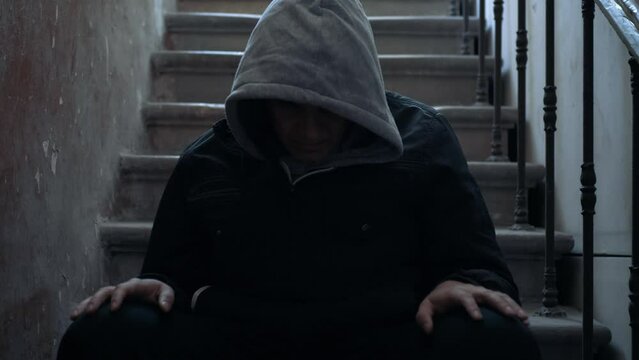 Lonely man in hood with his head down sits on stairs in dark. Stress and depression mental problems concept.