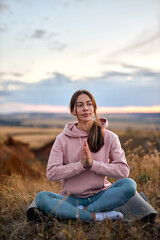 Fototapeta na wymiar pretty caucasian female keep calm, meditating on fitness mat sitting outdoors in nature, keep hands together, engaged in yoga. young lady in sportswear concentrated on mind, enjoying yoga