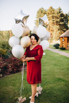 Beautiful smiling senior woman in red dress standing on green lawn with balloons. A woman celebrates her 50th years birthday. Beautiful woman at a party