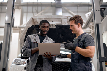 Black Car Mechanic And Caucasian Repairman Checking Diagnostics Results on Laptop, Computer. Explains an Engine Breakdown to Mechanic. Car Service Employees Inspect Car's Engine Bay. Clean Workshop.