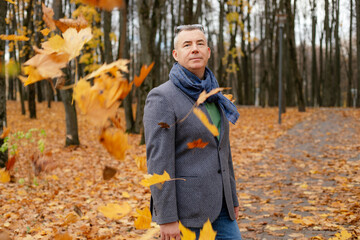 Smiling, confident, tranquil grizzled modern man in eyeglasses, warm clothes walking in gold park through fallen leaves