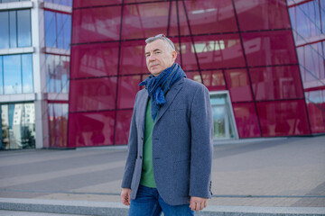 Confident and satisfied gray haired businessman in eyeglasses, wear jacket and scarf near red office building. Start up