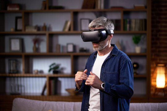 Adult Grey-haired Man Using Virtual Reality And Playing Video Games At Home. Joyful Man Using PlayStation And Competing In Virtual Games While Relaxing On Sofa