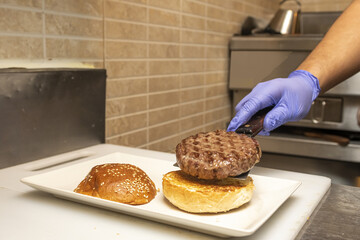 A chef with a gloved hand assembling a freshly cooked burger on top of a toasted muffin