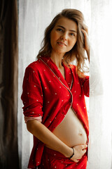 Vertical smiling, pensive pregnant blond woman holding naked belly, wear red pajamas, resting and...