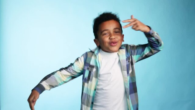 Cute energetic little african american kid boy dancing in good mood on isolated blue background Adorable kid in white t shirt and checked shirt having fun alone