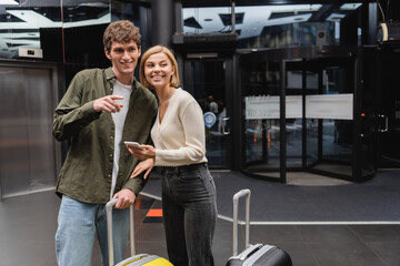young man pointing with finger and looking away near smiling girlfriend and travel bags in hotel.