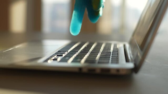 Close-up side view of unrecognizable man wearing protective gloves cleaning and removing dust from laptop keyboard using blue slimy absorbent gel, at table on background of sunny window, slow motion.
