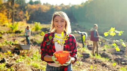 Portrait shot of blonde young beautiful Caucasian woman standing outdoor with tree seedling in pot and smiling cheerfully. Pretty female eco activist working against deforestation. Gardening. - Powered by Adobe
