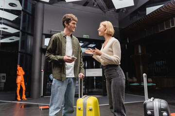 happy blonde woman talking to boyfriend with mobile phone near travel bags in hotel lobby.