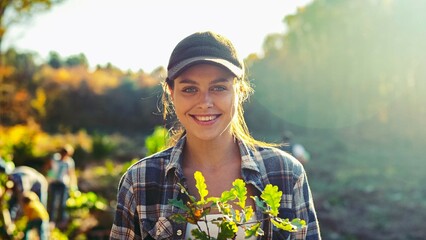 Portrait shot of young beautiful Caucasian woman standing outside with tree seedling in pot and smiling cheerfully.
