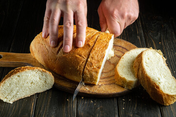 Cook's hand with a knife for cutting whole grain bread on a kitchen wooden board. Fresh bread on...