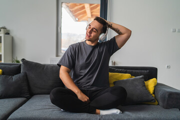 A young guy is sitting on couch meditating and stretching while listening to music and using his...