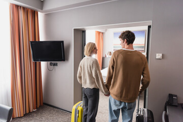 back view of young couple of travelers with suitcases holding hands and looking at each other in modern hotel suite.