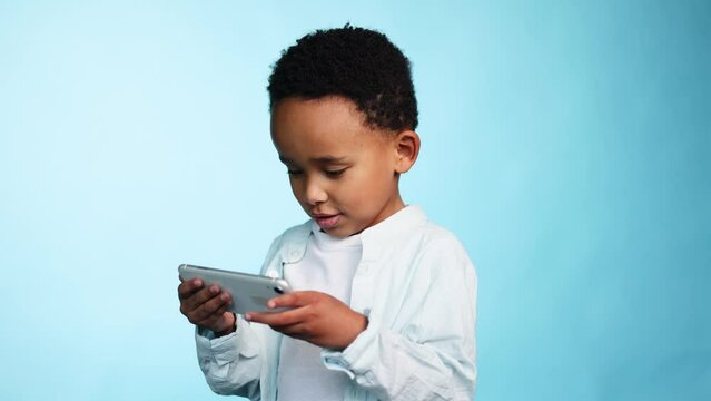 Adorable cute african american little boy child kid hold smartphone scrolling social media use educational children app application or play game on isolated blue background 