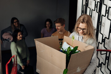 Smiling, glad, talking students moving in accommodation, taking out and carrying boxes with houseplant stuff on stairs