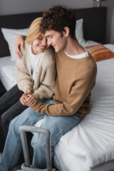 young couple of tourists holding hands while sitting with closed eyes in hotel bedroom.