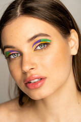 Brunette teen girl with multicolored makeup and freckles looking at camera isolated on grey.