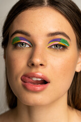 Teenage girl with colorful visage sticking out tongue and looking at camera isolated on grey.