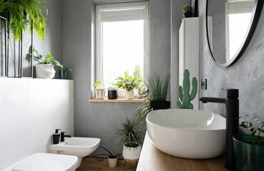 Stylish bathroom with window and green plants. Ceramic wash basin with tap, toilet, bidet and round mirror on the grey wall. New design in modern apartment.