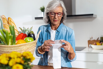 Lovely mature woman standing at home table holding grocery receipt discussing for rising prices.