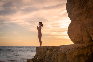 Woman standing on rocks by the sea practicing yoga at sunrise or setset.