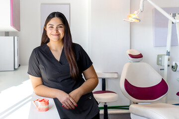 Mujer latina joven Dentista de pie recargada en consultorio Dental Sonriente  - obrazy, fototapety, plakaty