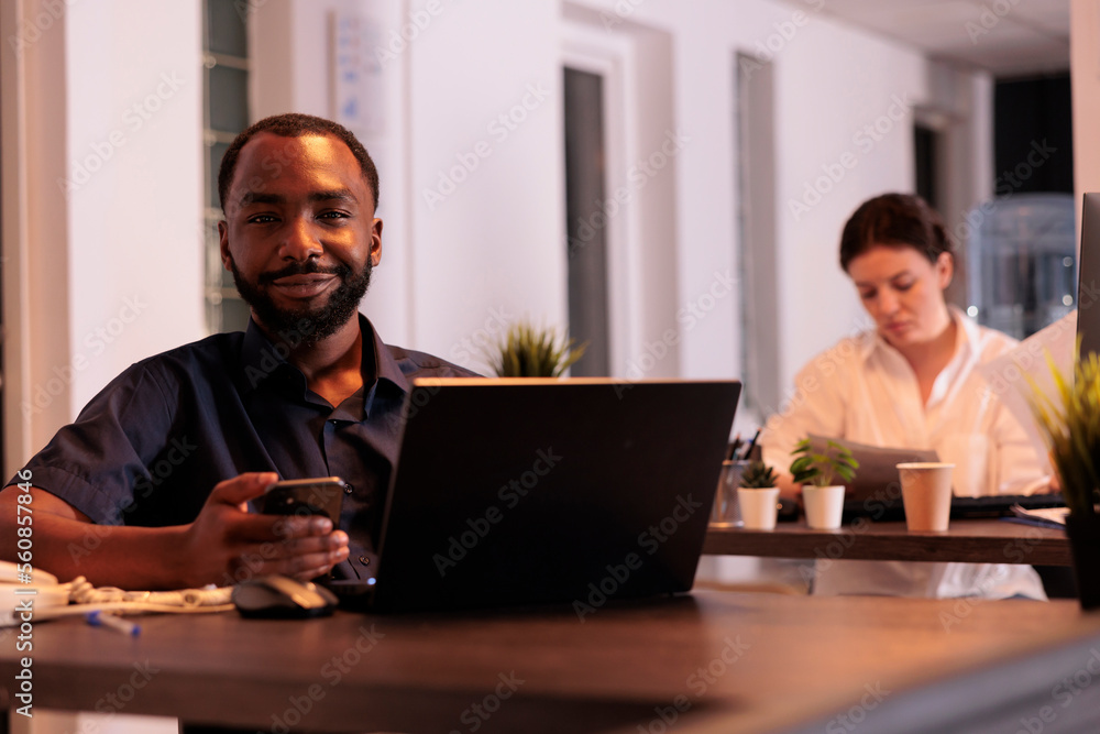 Poster smiling african american man working on startup project on laptop, using smartphone appplication in 