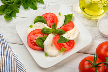  Caprese salad with mozzarella cheese, tomatoes and basil in a square white plate on a white rustic table. - 560858826