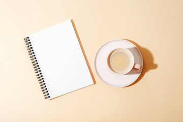 Obraz na płótnie Canvas Coffee in cup and spiral notebook on neutral background with sharp shadows. Top view, flat lay, mockup