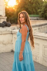Naklejka premium Woman sunset blue dress. Portrait of a woman with long hair and a blue dress against the backdrop of the setting sun and a white building. Lifestyle, walking around the city