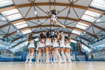 Foto op Plexiglas A group of cheerleaders are captured in a dynamic and athletic moment, performing a stunt by lifting one of their team members into the air. Showcasing energy, athleticism and unity in cheerleading. © PoppyPix