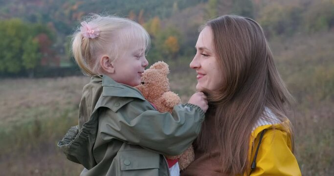 Young woman has fun with her fair-haired child in nature. Happy mother hugs and kisses her little daughter.