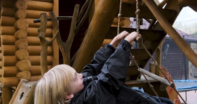 Little blond boy laughs and tries to climb rope ladder. Child actively playing in wooden playground outdoor
