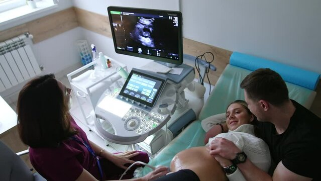 Happy smiling expecting couple having a baby ultrasound check. Pregnant lady lies on her back and husband supports her. Top view.