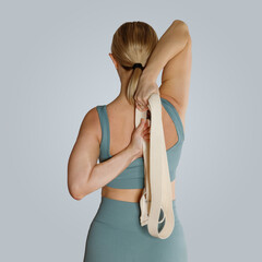 Fitness young woman doing yoga exercise in sportswear with strap in studio, back view. Pilates and stretching concept