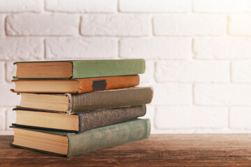 Stack of old hardcover books on wooden table near white brick wall, space for text