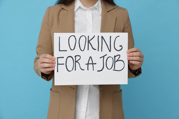 Young unemployed woman holding sign with phrase Looking For A Job on light blue background, closeup
